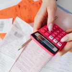 Mãos usando uma calculadora rosa para organizar despesas entre recibos e documentos financeiros.
