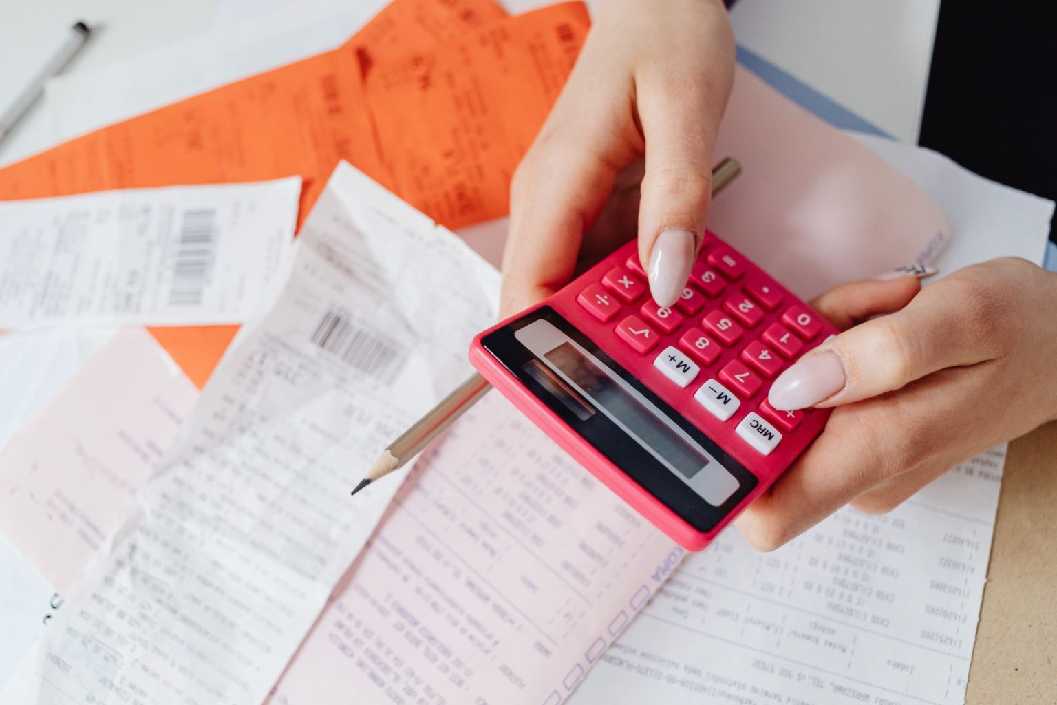 Mãos usando uma calculadora rosa para organizar despesas entre recibos e documentos financeiros.