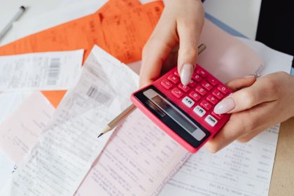 Mãos usando uma calculadora rosa para organizar despesas entre recibos e documentos financeiros.