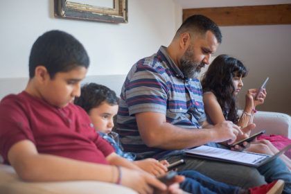 Pai e três crianças sentados em um sofá usando dispositivos eletrônicos em casa.